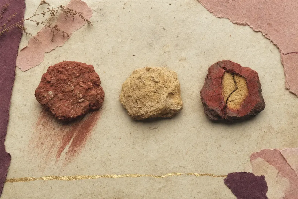 Three lumps of coloured earth in a row: a gritty red one streaked across a stone, a yellow one, and a split piece with a deep brick-red outer rind and a still-yellow centre.