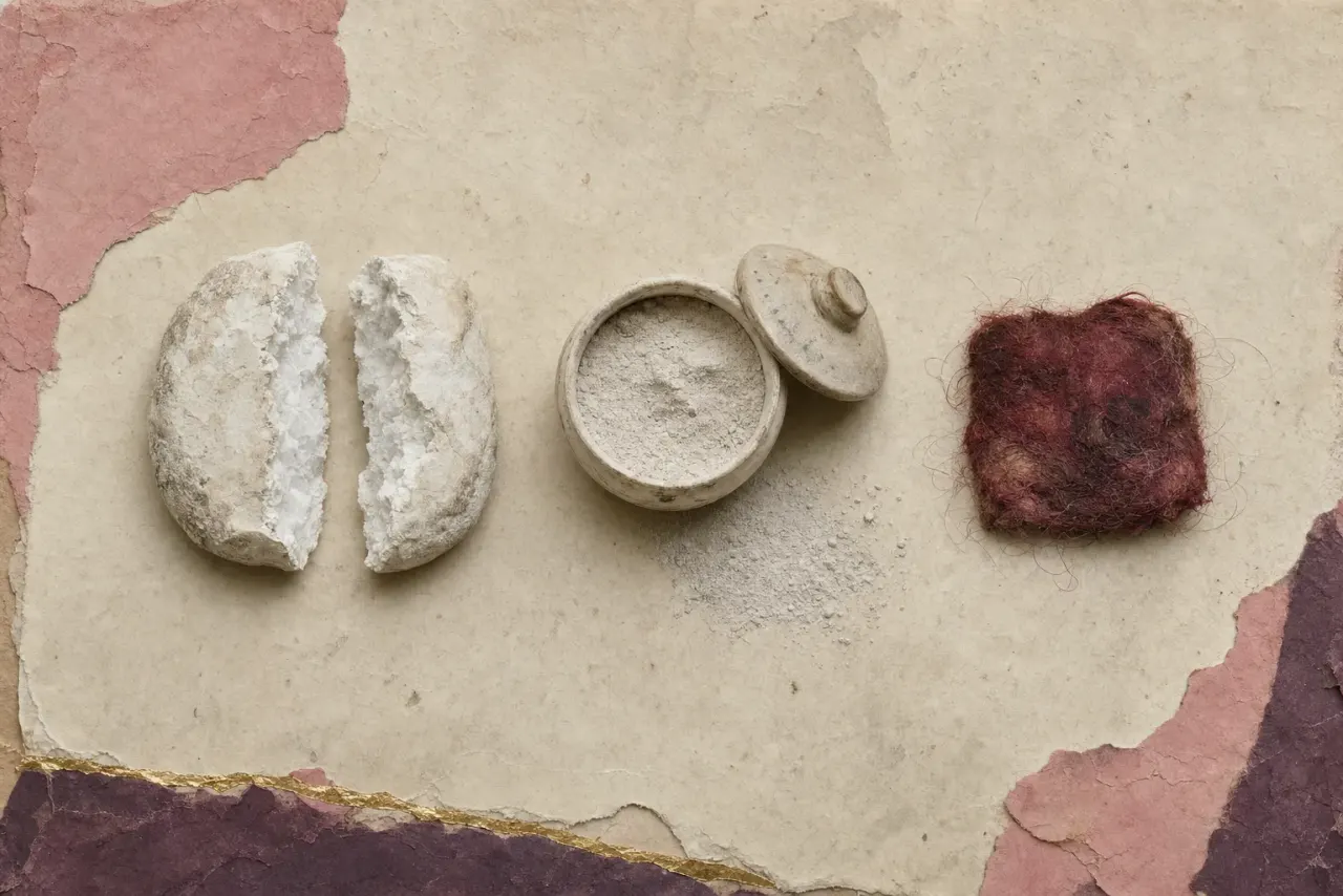 A cake of dense white lead pigment broken open to show its crystalline interior, beside a lidded jar of the ground powder and a small pad of hair loaded with red.