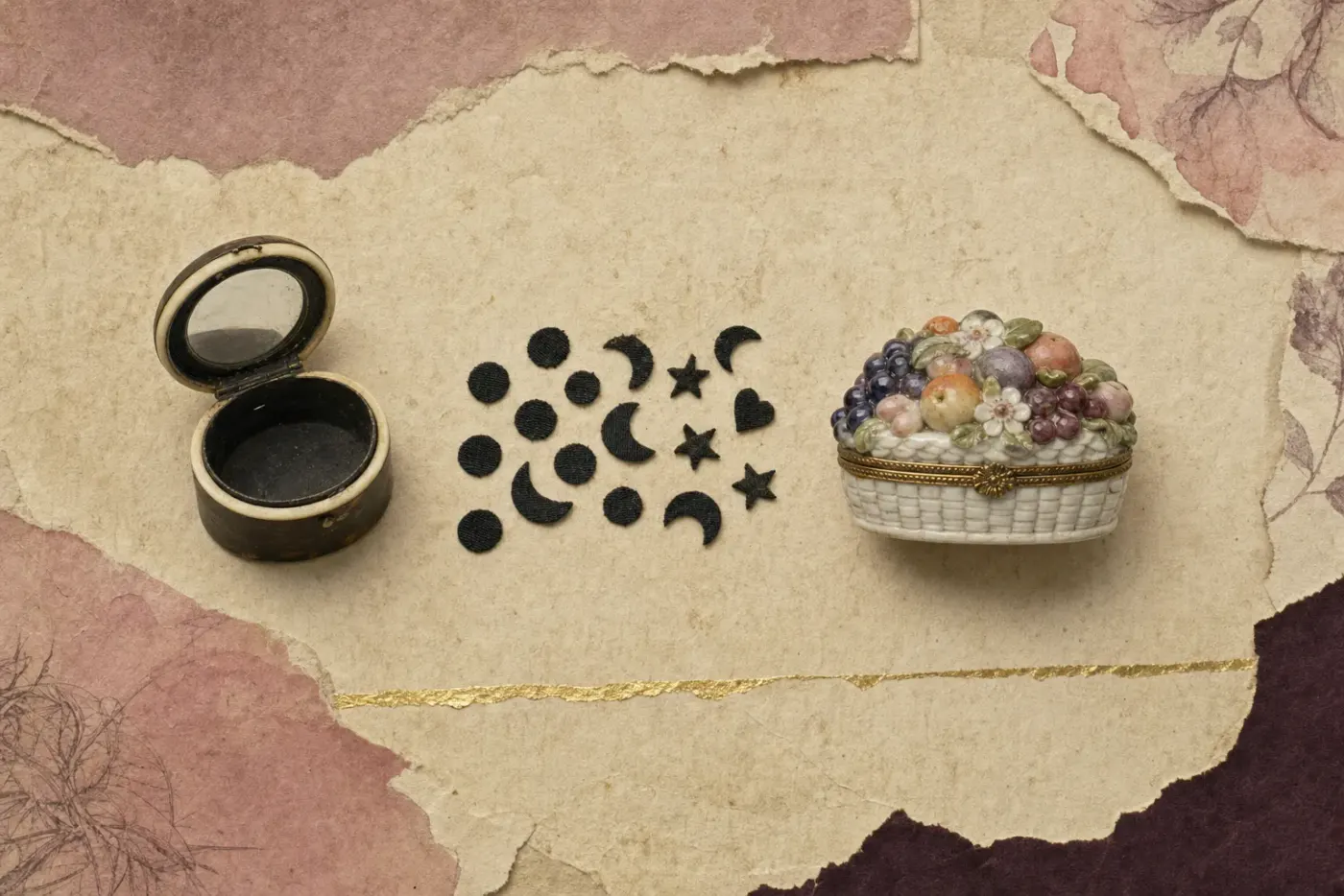 The makings of an eighteenth-century face patch: a small round box of dark horn with an ivory rim, open to show a mirror inside the lid; a scatter of tiny black patches cut as dots, crescents, stars and one heart; and a porcelain box modelled as a basket heaped with fruit and flowers, with a gilt mount.