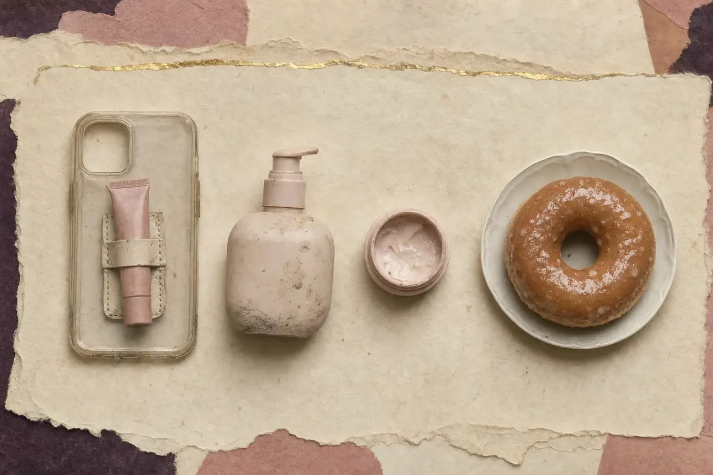 A clear phone case with a lip-treatment tube clipped to its back, a pump bottle of serum, a small pot of cream and a glazed doughnut on a white plate.