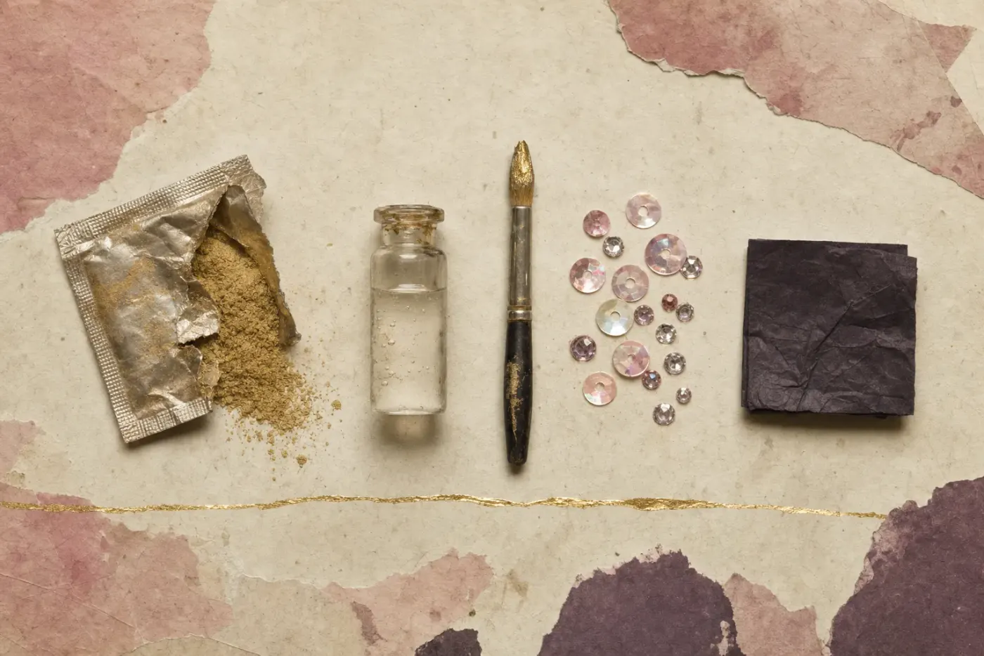 A torn sachet spilling fine gold pigment beside a vial of clear liquid, a brush tipped with liquid gold, scattered sequins and rhinestones, and a folded square of black tissue.