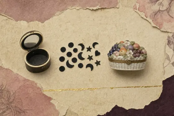 The makings of an eighteenth-century face patch: a small round box of dark horn with an ivory rim, open to show a mirror inside the lid; a scatter of tiny black patches cut as dots, crescents, stars and one heart; and a porcelain box modelled as a basket heaped with fruit and flowers, with a gilt mount.