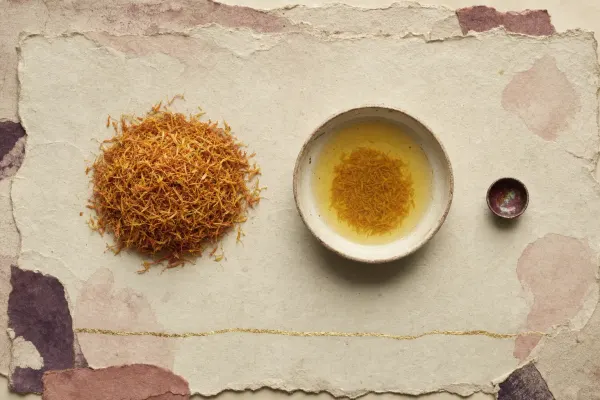 A large heap of dried orange-yellow safflower petals beside a bowl of yellow wash water and a very small cup painted inside with a thin film of deep crimson showing green iridescence.