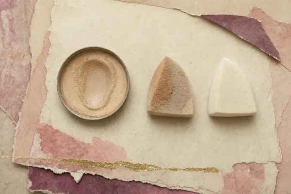 A shallow round pan of compacted cream-beige cake foundation, its surface swept into a shallow trough by use, laid beside a damp wedge sponge stained with the same beige and a second clean dry sponge.