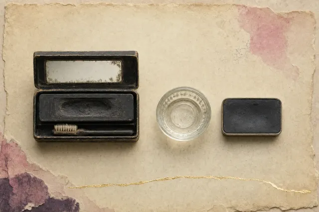 A cake mascara kit of the early twentieth century: a small black box lying open with a hard black cake worn hollow in the middle, a tiny stiff brush beside it and a mirror in the lid, next to a small glass dish of water and a second black cake in a thin metal tray.