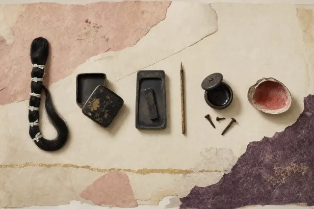 The things of a Heian court lady in a row: a long switch of black hair tied with paper cord, a lacquer box, an ink stone and brush, a bowl of dark tooth dye with rusted nails, and a shell painted inside with safflower red.