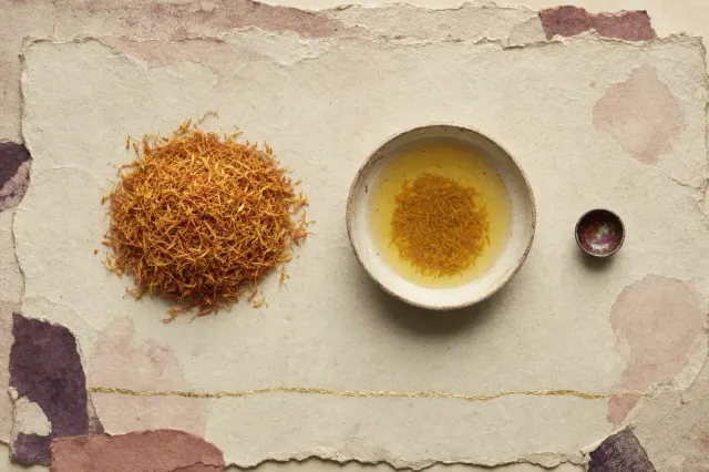 A large heap of dried orange-yellow safflower petals beside a bowl of yellow wash water and a very small cup painted inside with a thin film of deep crimson showing green iridescence.