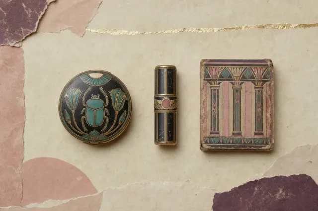 Mid-1920s cosmetic packaging in the Egyptian revival style: an enamelled compact with a scarab and lotus lid, a lipstick case banded with a winged disc, and a powder box printed with stylised papyrus columns.
