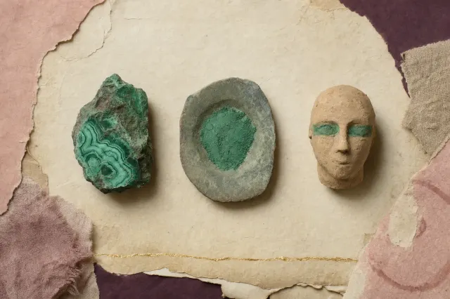 A lump of raw green malachite ore, a worn siltstone grinding palette holding a crust of the same green, and a small modelled clay head with green pigment around both eyes, laid out in a row.