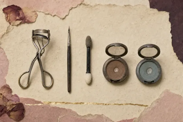 A metal eyelash curler, a fine pointed liner brush, a sponge-tipped applicator and two small shallow compacts of pressed eyeshadow laid out in a row.