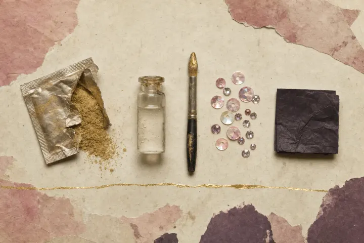 A torn sachet spilling fine gold pigment beside a vial of clear liquid, a brush tipped with liquid gold, scattered sequins and rhinestones, and a folded square of black tissue.