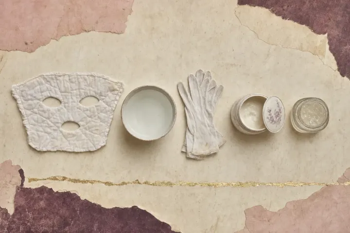 The night mask across centuries: a quilted white cotton face mask with holes for the eyes and mouth, a white bowl of water, a pair of white cotton gloves, a porcelain pot of cold cream with a flowered lid and a glass jar of clear petroleum jelly.