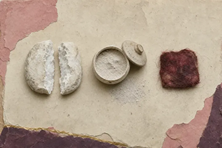 A cake of dense white lead pigment broken open to show its crystalline interior, beside a lidded jar of the ground powder and a small pad of hair loaded with red.
