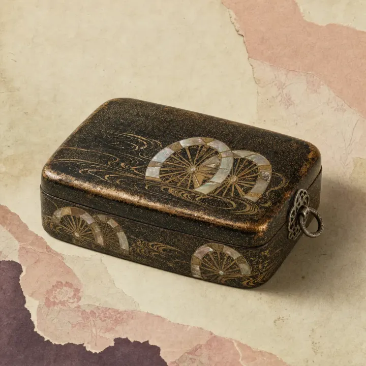 A Japanese lacquered cosmetic box with rounded corners and a fitted lid, its black surface flecked with gold and decorated with ox-cart wheels half sunk in swirling water, the wheels inlaid in mother-of-pearl, with an openwork silver fitting and ring on the side.